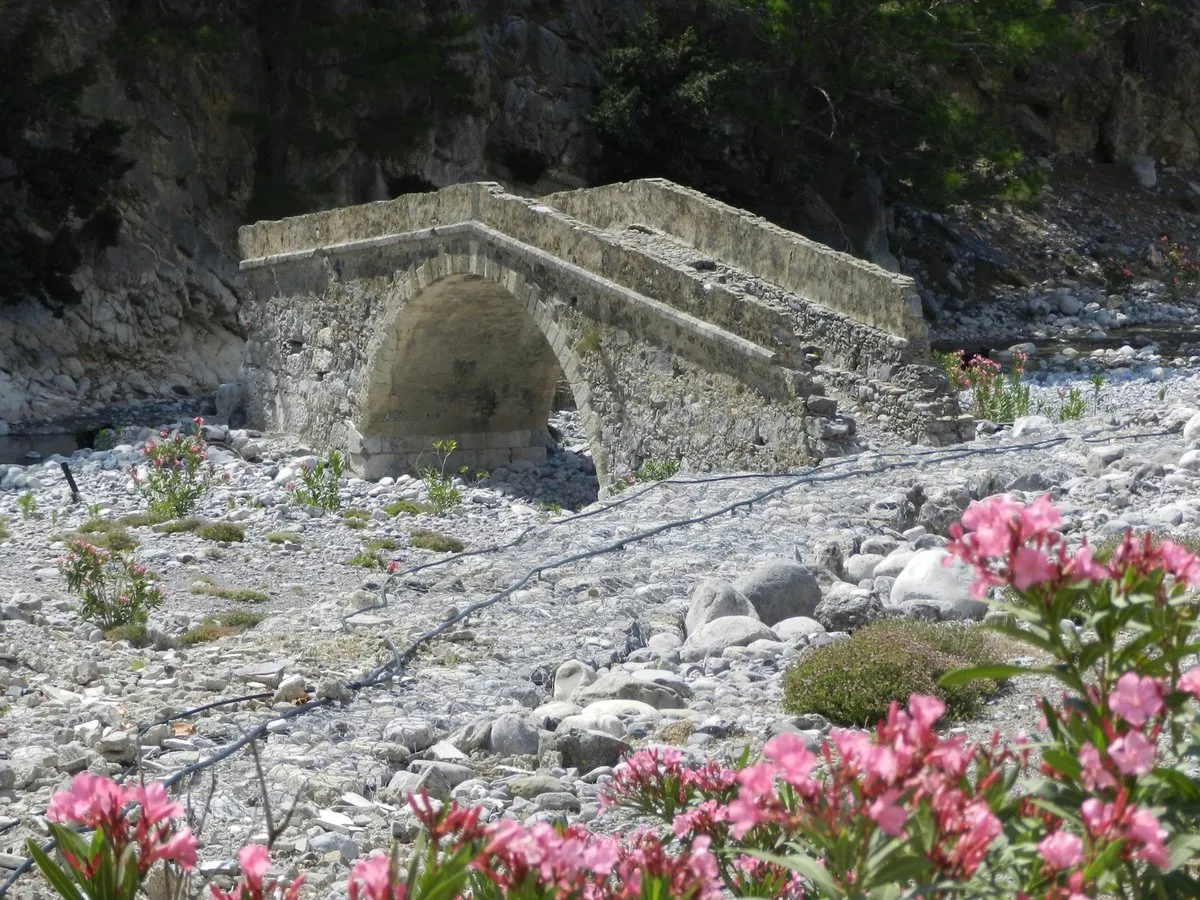 Eine alte steinerne Bogenbrücke über ein trockenes Flussbett aus weißen Steinen, im Vordergrund Oleander