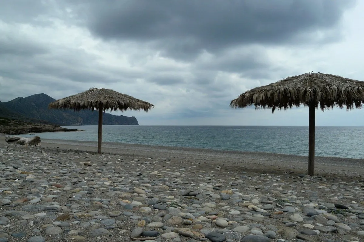 Der Strand Gialiskari im Winter: graue Kiesel, zwei Strohschirme und ein bewölkter Himmel über dem Libyschen Meer