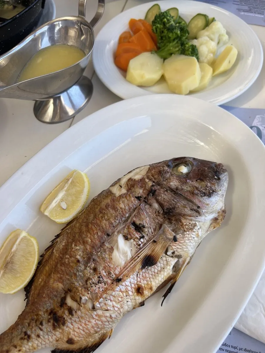 A whole grilled fish with lemon wedges, a jug of ladolemono and boiled greens on a taverna table