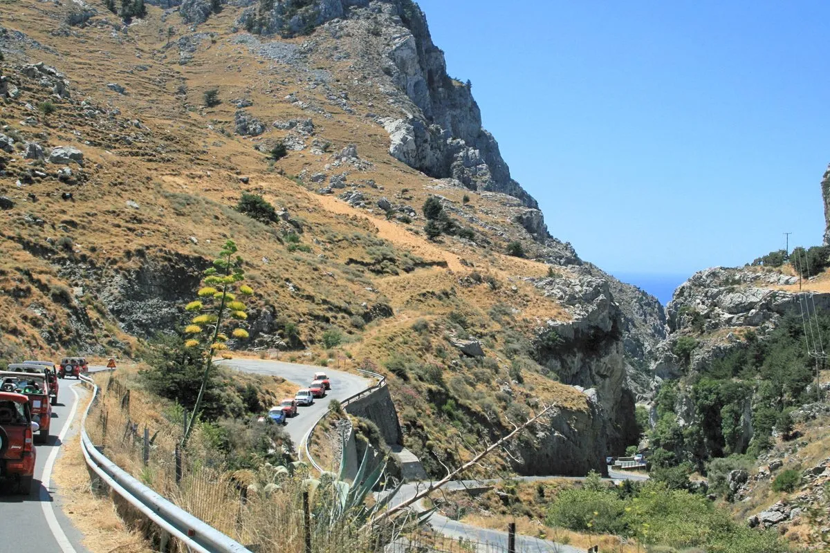 Die Straße windet sich zwischen trockenen Hängen hinab, mit einem Blick aufs Meer