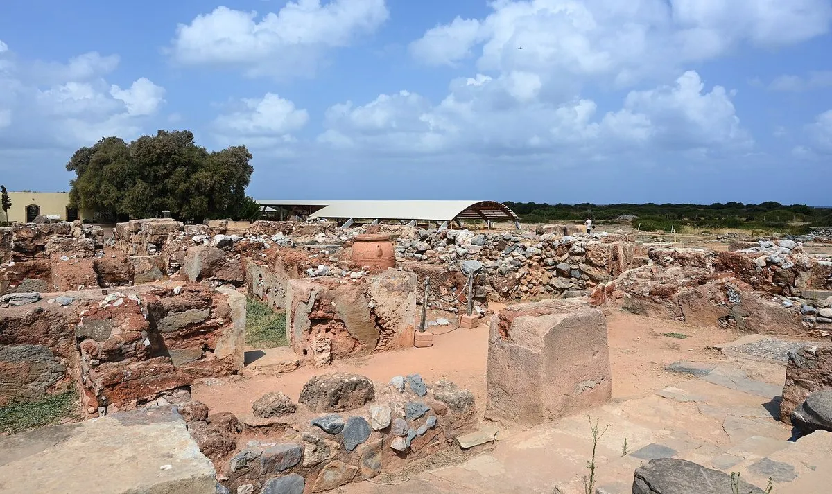 The excavated ruins of the Minoan palace at Malia, low stone walls across a flat site with the modern shelter roof and the sea behind