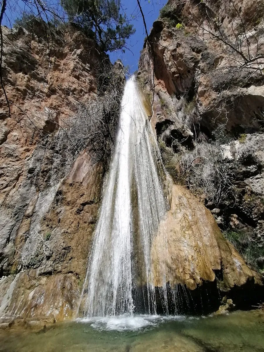 Der Mylonas-Wasserfall stürzt als einzelne weiße Säule eine orangefarbene Felswand hinab in ein grünes Becken